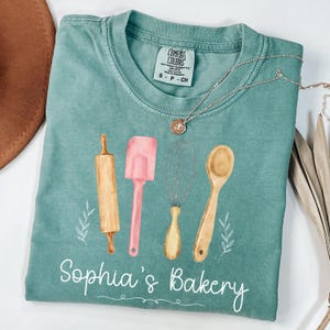 Peut inclure: Un t-shirt Comfort Colors turquoise avec un motif aquarelle d'ustensiles de cuisine : un rouleau &agrave; p&acirc;tisserie, une spatule, un fouet et une cuill&egrave;re en bois. Le texte "Sophia's Bakery" est &eacute;crit en blanc en dessous. Un collier argent&eacute; avec un pendentif est pos&eacute; sur le t-shirt.