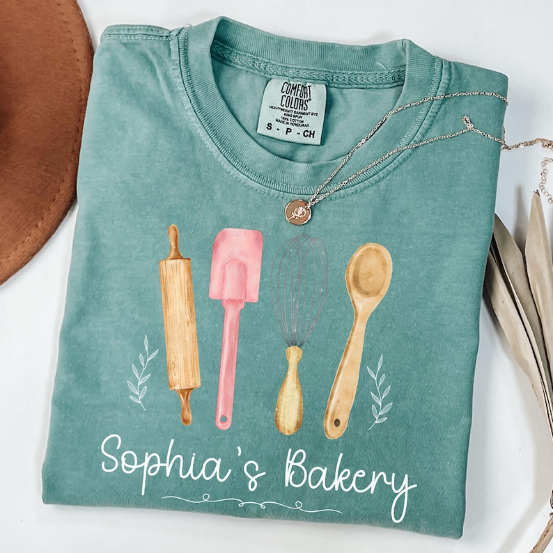 Peut inclure: Un t-shirt Comfort Colors turquoise avec un motif aquarelle d'ustensiles de cuisine : un rouleau &agrave; p&acirc;tisserie, une spatule, un fouet et une cuill&egrave;re en bois. Le texte "Sophia's Bakery" est &eacute;crit en blanc en dessous. Un collier argent&eacute; avec un pendentif est pos&eacute; sur le t-shirt.