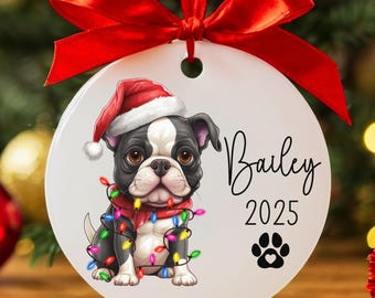 May include: White round Christmas ornament with a cartoon image of a black and white Boston Terrier wearing a Santa hat and a red scarf with colorful Christmas lights wrapped around it. The ornament has the text "Bailey 2025" and a paw print with a heart.
