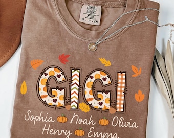 May include: Brown Comfort Colors t-shirt with the name "GIGI" in a fall-themed design, including pumpkins and leaves. The names Sophia, Noah, Olivia, Henry, and Emma are printed below. A silver necklace rests on the shirt.