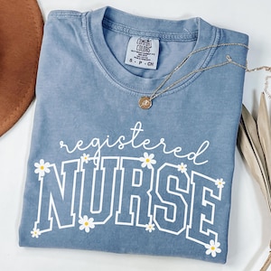 May include: A blue Comfort Colors t-shirt with the words "registered NURSE" in white, decorated with small daisy graphics. A gold necklace with a pendant rests on the shirt.