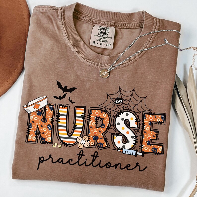 May include: A brown Comfort Colors t-shirt with a Halloween-themed design. The shirt features the words "NURSE practitioner" in a spooky font with bats, a spider web, and medical-themed graphics. A silver necklace rests on the shirt.