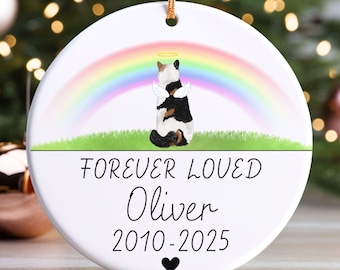May include: A white ceramic Christmas ornament with a gold string for hanging. The ornament features a colorful rainbow, a cartoon cat with angel wings and a halo, and the text "FOREVER LOVED Oliver 2010-2025" with a heart.