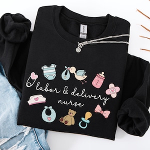 May include: Black sweatshirt with a white graphic design that says "labor & delivery nurse" and includes baby-related icons like a pacifier, a baby bottle, a onesie, a teddy bear, and a safety pin.