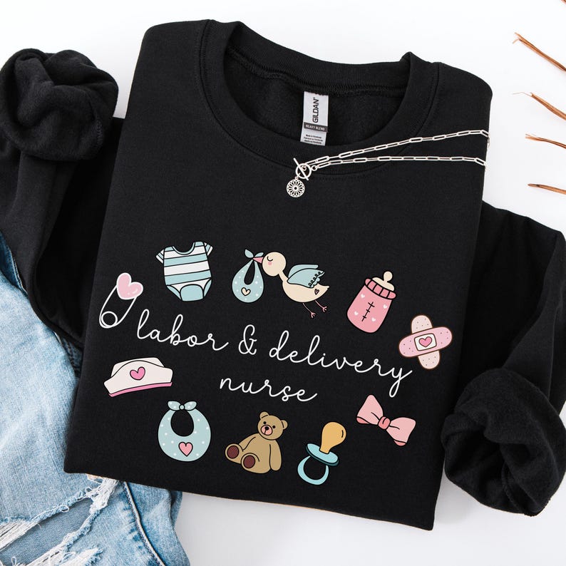 May include: Black sweatshirt with a white graphic design that says "labor & delivery nurse" and includes baby-related icons like a pacifier, a baby bottle, a onesie, a teddy bear, and a safety pin.