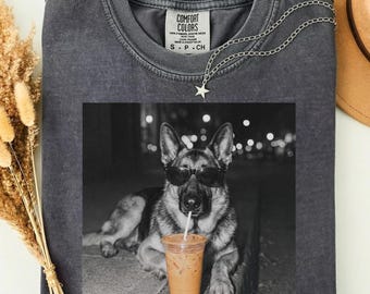 May include: A charcoal gray crew neck t-shirt featuring a black and white graphic of a German Shepherd dog wearing sunglasses and holding a straw in a plastic cup. The shirt has a Comfort Colors label at the inner neckline and a star charm necklace.