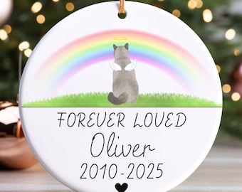 May include: A white ceramic ornament with a gold string for hanging. The ornament features a gray cat with angel wings and a halo, sitting under a rainbow. Text reads "FOREVER LOVED" and "Oliver 2010-2025" with a heart.