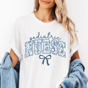 May include: White t-shirt with a blue and white floral design that says "pediatric NURSE" with a blue bow underneath.