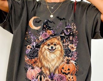 May include: A charcoal gray t-shirt featuring a graphic print of a fluffy Pomeranian dog wearing a witch hat. The design includes a crescent moon, bats, a haunted house, orange pumpkins, and various colorful flowers surrounding the dog.
