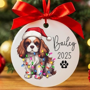 May include: A white round Christmas ornament with a cartoon image of a Cavalier King Charles Spaniel wearing a Santa hat and wrapped in Christmas lights. The ornament is personalized with the name "Bailey" and the year "2025".