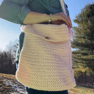 May include: A light pink crochet shoulder bag with a flap closure and a wooden button. The bag has a thick, textured stitch and a matching strap. The background shows a sunny outdoor scene.