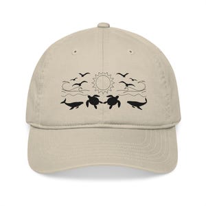 May include: A beige baseball cap with a black graphic depicting a sun, birds, waves, whales, and sea turtles. The cap has a curved peak and a fabric strap closure. This hat is a great accessory for anyone who loves the ocean.
