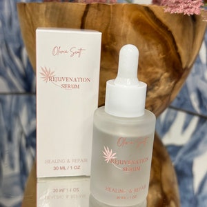 May include: A frosted glass serum bottle and box with the text "Olivia Scott Rejuvenation Serum" in pink. The bottle has a dropper and the box and bottle both read "Healing & Repair 30 ml / 1 oz". The background includes a wooden bowl and pink flowers.