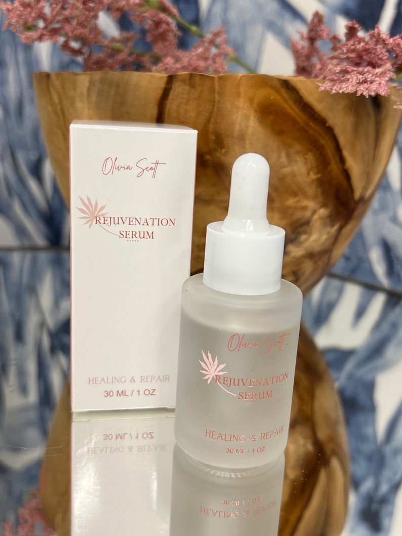 May include: A frosted glass serum bottle and box with the text "Olivia Scott Rejuvenation Serum" in pink. The bottle has a dropper and the box and bottle both read "Healing & Repair 30 ml / 1 oz". The background includes a wooden bowl and pink flowers.
