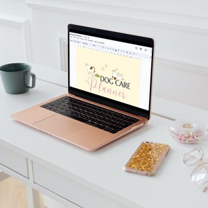May include: A rose gold laptop computer open to a Google Docs document with the text "DOG CARE PLANNER" in pink and white lettering. The document has a cartoon image of two dogs. The laptop is on a white desk with a white mug, a clear container with pink and gold pills, a gold glitter phone case, and clear glasses.