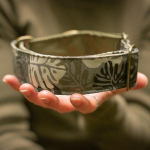 Op de afbeelding: Een hondenhalsband met een bladerpatroon in grijstinten en olijfgroen. De halsband heeft een messingkleurige metalen gesp en D-ring. De stof heeft een herhalend patroon van gestileerde bladeren. De halsband wordt in een hand gehouden.