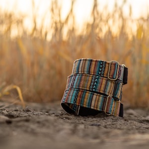 May include: Three colorful dog collars stacked on top of each other. The collars are made of woven fabric with a geometric pattern in shades of blue, orange, yellow, and brown. The collars have silver metal buckles.