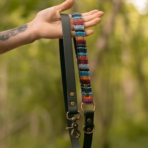 May include: A black leather dog lead with a colourful woven fabric section. The lead has brass-coloured hardware, including a clasp and D-rings. The woven fabric has a geometric pattern with red, blue, orange, and white colours.