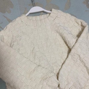Könnte beinhalten: Ein cremefarbener Strickpullover mit Zopfmuster. Der Pullover hängt an einem weißen Kleiderbügel.