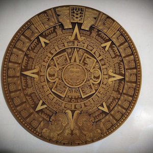 Puede incluir: Una placa de pared circular de madera con un relieve detallado cortado con láser de la Piedra del Sol azteca. El diseño incluye un rostro central rodeado de anillos concéntricos con patrones geométricos complejos y glifos.