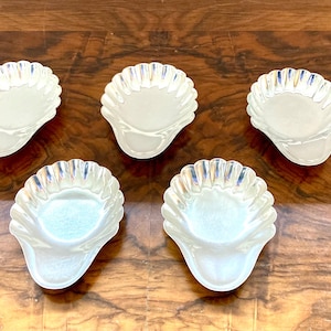 May include: Five iridescent, shell-shaped dishes are arranged on a wooden surface. The dishes have a scalloped edge and a pearlescent finish, reflecting light with hints of blue and gold. The overall aesthetic is vintage and decorative.