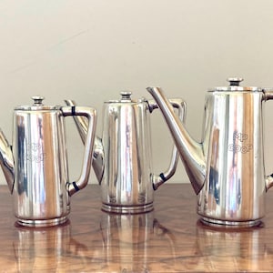 Könnte beinhalten: Drei silberfarbene Teekannen mit Ausgüssen und Griffen stehen auf einer Holzoberfläche. Die Teekannen haben eine glänzende, reflektierende Oberfläche. Die mittlere Teekanne ist kürzer als die anderen beiden. Jede Teekanne trägt die Initialen "MP COOP".