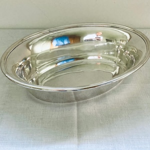 Op de afbeelding: Een glanzende, ovale zilveren serveerschaal. De schaal heeft een glad, reflecterend oppervlak en een licht verhoogde rand. Hij staat op een wit tafelkleed en weerspiegelt de omgeving. De schaal wordt waarschijnlijk gebruikt om voedsel te serveren.