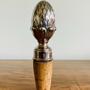 May include: A silver-colored bottle stopper with a detailed, artichoke-like design. The stopper has a cork base and a metallic top, suitable for wine bottles. The design is intricate and decorative.