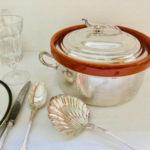 Puede incluir: Una fuente de servir de plata con un borde de cerámica marrón y un asa de lagarto de plata en la tapa. La fuente está sobre una superficie blanca con una cuchara y un cuchillo de plata.