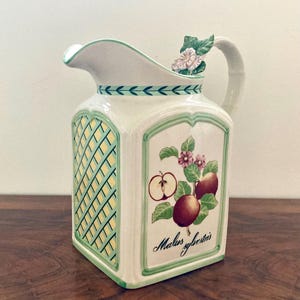 Könnte beinhalten: Weißer Keramikkrug mit quadratischem Körper und gebogenem Ausguss. Der Krug hat auf einer Seite ein grün-gelbes Gittermuster und auf der anderen eine Apfelillustration mit dem Text "Malus sylvestris". Der Griff ist mit einem floralen Akzent verziert.