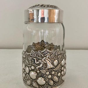 Puede incluir: Un tarro de cristal transparente con tapa plateada y una banda decorativa plateada. La banda presenta un diseño en relieve detallado de frutas, hojas y figuras. El tarro se utiliza probablemente para almacenamiento o exhibición.