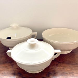 Puede incluir: Tres cuencos de servir de cerámica blanca con tapas. Dos de los cuencos tienen tapas con asas. Los cuencos están sobre una superficie de madera.