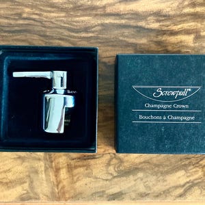 May include: A chrome champagne crown opener in a black box. The box is open, revealing the opener. The box next to it has the text "Screwpull Champagne Crown Bouchons à Champagne".