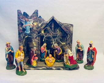 May include: A detailed nativity scene with figures of Mary, Joseph, baby Jesus, the Three Wise Men, and other biblical characters. The scene includes a rustic stable with string lights and an angel. The figurines are painted in vibrant colors.