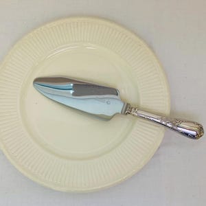 Op de afbeelding: Een zilveren taartschep ligt op een crèmekleurige bord met een geribbeld ontwerp. De schep heeft een glanzend blad en een decoratief handvat. Het bord en de schep staan tegen een witte achtergrond.