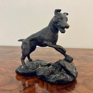 May include: A bronze sculpture of a dog standing on a rocky base. The dog is in a playful pose, with its head tilted and its mouth slightly open. The sculpture is detailed and realistic, with a smooth, dark brown finish.