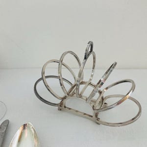 Puede incluir: Porta servilletas plateado con un diseño en forma de abanico. El porta servilletas está hecho de múltiples anillos entrelazados.