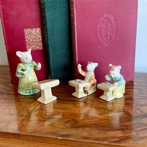 Op de afbeelding: Een verzameling vintage keramische muisbeeldjes in een klaslokaal. Eén muis houdt een boek vast, terwijl anderen aan bureaus zitten. De beeldjes staan tegen een achtergrond van antieke boeken met rode en groene kaften.