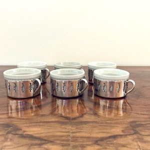 May include: Six silver cups with white interiors and small handles are displayed on a wooden surface. The cups feature a decorative pattern of small cut-out shapes around the base. These cups are likely for serving beverages.