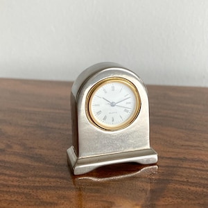 Può includere: Un orologio da tavolo argentato con parte superiore ad arco e base rettangolare. Il quadrante è bianco con numeri romani neri, lancette nere sottili e una lunetta circolare dorata. La parola Quartz è stampata in nero sul quadrante.