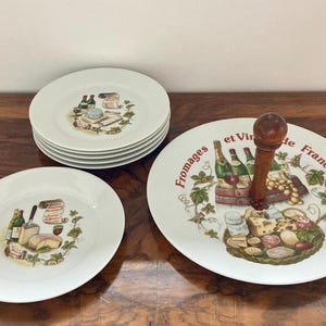 Op de afbeelding: Een witte keramische kaas- en wijnservies. De set bevat een etagère met een houten handvat, verschillende borden en een kleiner bord. Elk stuk heeft een kleurrijke illustratie van kaas, wijnflessen en druiven.