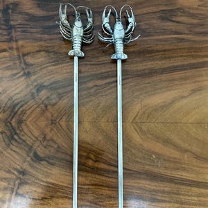 May include: Two silver-colored lobster skewers. Each skewer features a detailed lobster figure at the top and a long, pointed shaft. The skewers are displayed on a polished, dark wood surface.