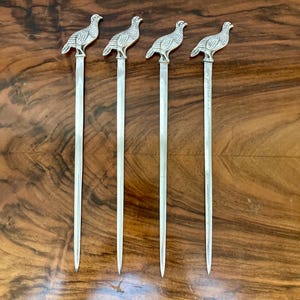 May include: Four silver cocktail picks, each topped with a detailed bird figure. The picks have long, slender stems and are arranged on a dark wooden surface. The birds are facing left.