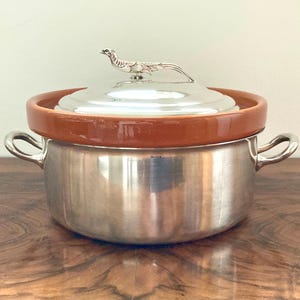 Puede incluir: Una olla de plata con una tapa de terracota marrón y un pomo de plata en forma de faisán. La olla tiene dos asas y está sobre una superficie de madera.