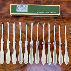 May include: A set of twelve silver-toned fruit forks with a three-pronged design. The forks have a teardrop-shaped handle with vertical lines and a decorative element. A green box with the text "Gense" is in the background.