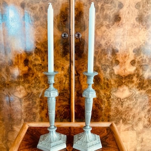 May include: Two matching silver-colored candle holders with white candles. The candle holders have a detailed design and are placed on a wooden surface. The background is a wooden cabinet.