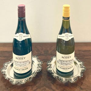 May include: Two bottles of Antonin Rodet wine, one Pinot Noir and one Chardonnay, sit on ornate silver trays. The Pinot Noir bottle has a red cap, while the Chardonnay has a yellow cap. Both bottles have white labels.