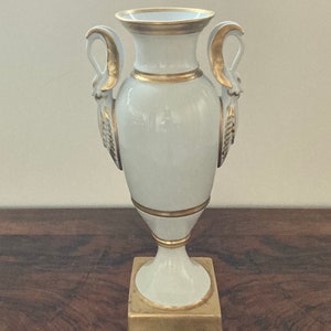 Puede incluir: Jarrón de porcelana blanca con detalles dorados y asas en forma de cisne. El jarrón tiene una base cuadrada y una forma de urna clásica.