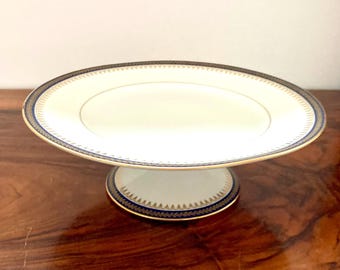 Puede incluir: Un soporte para pastel blanco con un borde azul y dorado. El soporte tiene una base redonda y una parte superior circular.