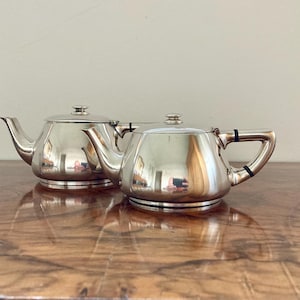 Op de afbeelding: Twee gepolijste zilveren theepotten met deksels, op een houten oppervlak. De theepotten hebben gebogen tuiten en handvatten, die de omgeving weerspiegelen. De theepotten zijn een klassiek ontwerp, voor het serveren van thee.